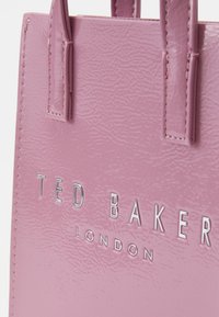 Vaaleanpunainen, kiiltävä kassi teksturoidulla pinnalla, jossa on kohokuvioitu hopeinen "TED BAKER LONDON" -logo ja tukevat kahvat.