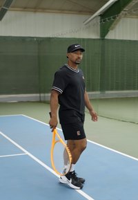Černé sportovní tričko s bílým lemem, černé kraťasy, držící oranžovou tenisovou raketu, na modrém vnitřním kurtu s zeleným sítí v pozadí.
