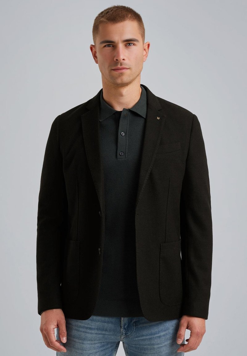 Zwarte blazer van gestructureerde stof, met enkele knoopsluiting en twee voorzakken, gedragen over een donker polo-shirt, aangevuld met lichtblauwe jeans.