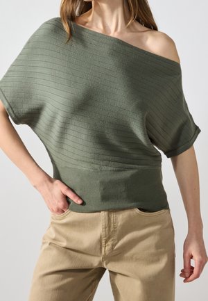 Vrouw draagt een geribd olijfgroen off-shoulder top met korte mouwen, ingestopt in beige broek, met één hand in de zak.