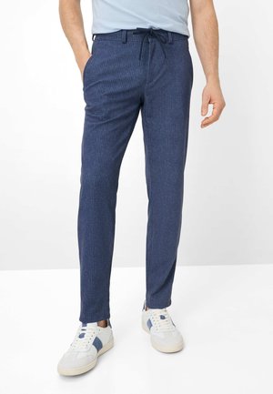 Uomo che indossa pantaloni con coulisse a righe sottili blu navy e scarpe da ginnastica bianche con accenti blu, in piedi davanti a uno sfondo semplice.