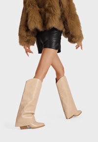 Botas de caña alta color beige con un acabado suave, con tacón cuadrado y un ligero pliegue en la parte superior, combinadas con una chaqueta de piel marrón y pantalones cortos negros.