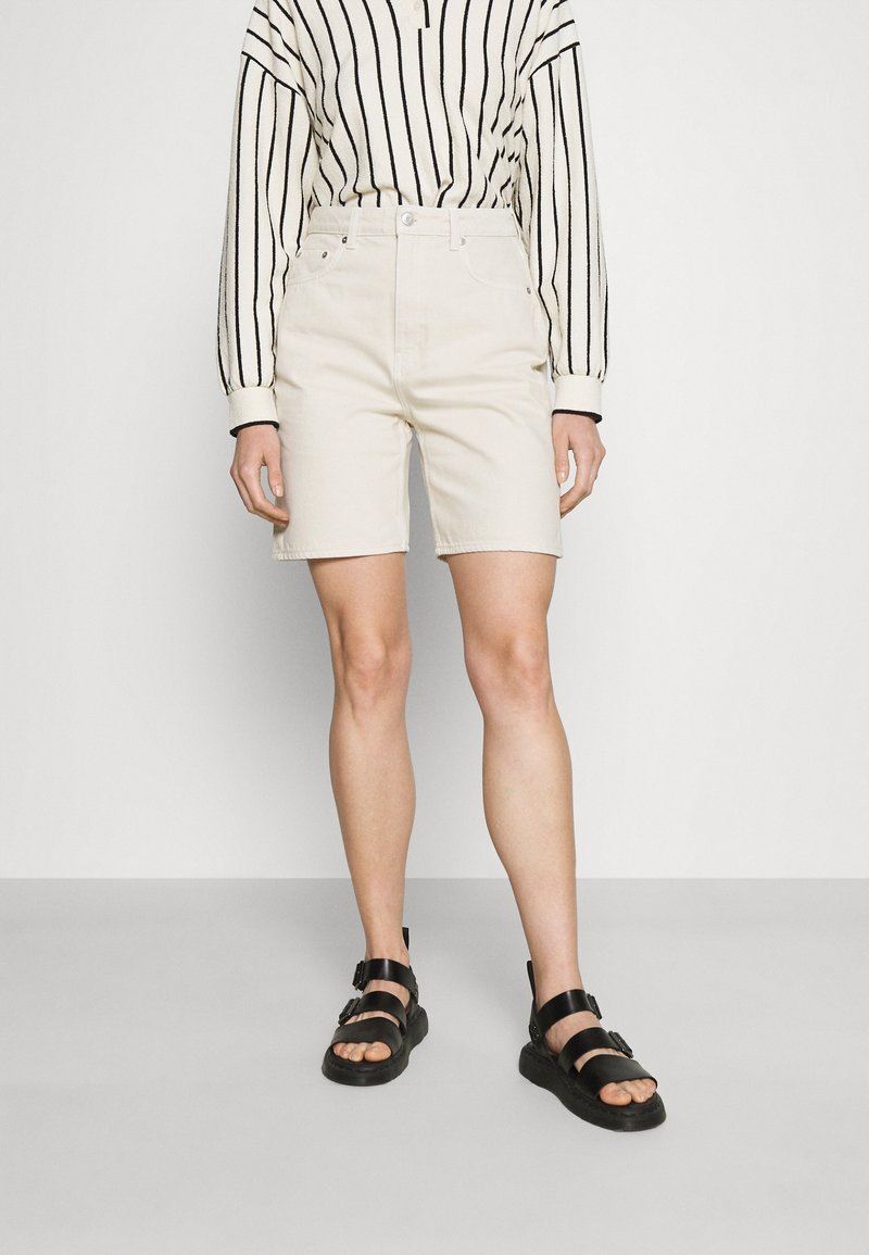 Personne portant un short crème taille haute, des sandales noires à brides et un haut blanc à manches longues avec des rayures verticales noires.