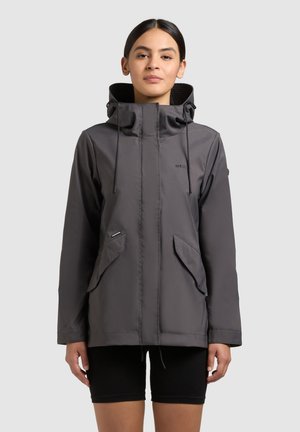 Naine, kes kannab tumehalli kapuutsiga jope ees taskutega ja musti lühikesi pükse, seisab ühevärvilise heledahalli tausta ees.