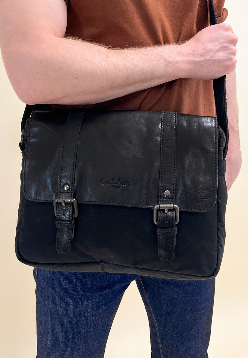 Juodas odinis paštininko krepšys su lygiu paviršiumi, dviem priekiniais sagtimis ir viršutine flip. Jame yra reguliuojama juosta ir stačiakampio formos.