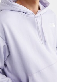 Sudadera de color púrpura claro hecha de una tela suave, con un bolsillo tipo canguro, capucha con cordón y un sutil detalle de logo en el pecho.