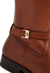 Brune skinnstøvel med glatt overflate, med et gullfarget metallspenne som accent og strukturert sømdetalj på skaftet og rundt sålen.