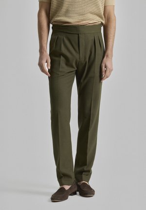 Pantalones ajustados en verde oliva con pliegues frontales, cinturilla plana y piernas ajustadas, combinados con mocasines marrones.