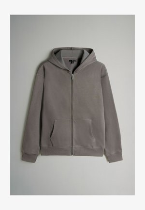 Šedá mikina na zip s přední kapsou, nastavovací kapucí, žebrovanými manžetami a hladkou texturou. Obsahuje jemné švy.