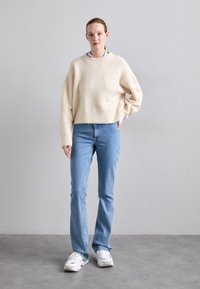 Maglione di lana lavorato a maglia chiaro con spalle scese, abbinato a jeans dritti azzurri. La modella indossa sneakers bianche, in piedi davanti a uno sfondo neutro.