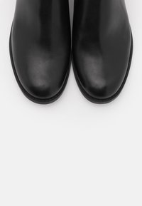 Par de botas de cuero negro con punteras redondeadas, vistas desde arriba sobre un fondo gris claro.