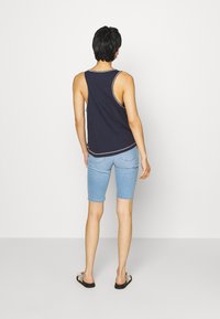 Marineblauwe tanktop met contrasterende stiksels, gecombineerd met lichtblauwe denim shorts. Model draagt zwarte sandalen, gezien van achteren tegen een effen achtergrond.