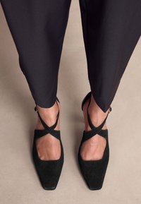 Czarne zamszowe szpilki z kwadratowym noskiem i paskiem krzyżowym. Charakteryzują się gładkim wykończeniem oraz paskiem wokół kostki z ozdobnym elementem metalowym.