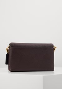 Tummanruskea nahkainen olkalaukku, jossa on teksturoitu pinta, kullanväriset metalliosat ja irrotettava ketjuhihna. Suorakulmainen muoto ja minimalistinen design.