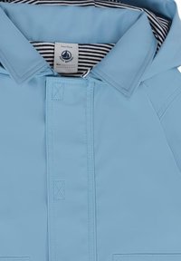 Giacca da pioggia azzurro chiaro con cappuccio, caratterizzata da un rivestimento interno a righe e una chiusura con bottoni semplice. Texture liscia e dettagli delle cuciture visibili.