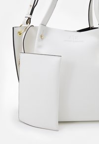 Bílá texturová kabelka vyrobená z umělého materiálu, s zlatými kovovými detaily, nastavitelnými popruhy a připojenou zipovou kapsou.