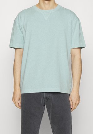 Mann trägt hellblaues T-Shirt mit kurzen Ärmeln und dunkelgraue Jeans, steht vor einem schlichten weißen Hintergrund.