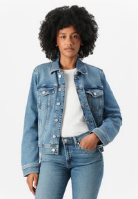 Jachetă din denim albastru deschis, cu un croi clasic, închidere cu nasturi, buzunare frontale și detalii cu cusături, asortată cu o cămașă albă și blugi.