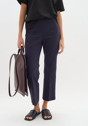 Marineblaue Hosen mit taillierter Passform und ausgestelltem Saum, kombiniert mit einem lockeren schwarzen T-Shirt und schwarzen Riemchensandalen, während sie eine braune Ledertasche hält.