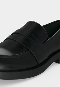 Μαύρο δερμάτινο loafer με χαμηλό τετράγωνο τακούνι και κλασική λεπτομέρεια λουριού στο πάνω μέρος, εμφανίζεται σε λευκό φόντο.