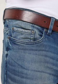 Vaqueros de mezclilla azul con textura desgastada, con un cinturón de cuero marrón. Los bolsillos cuentan con remaches de metal que acentúan el diseño clásico.