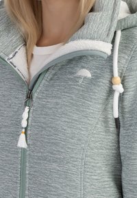 Ljusgrön zip-up hoodie med texturerad tyg, en plyschbeige foder och dekorativa dragsnören med trädetaljer. Subtilt paraplylogotyp.