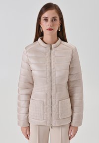Veste beige matelassée avec accents texturés, fermeture éclair et deux poches avant. Caractéristiques : coutures horizontales et col montant.