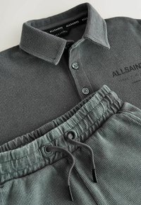 Sivé polo tričko s dlhým rukávom a vzorom waffle, s gombíkmi, v kombinácii so zodpovedajúcimi nohavicami so sťahovacou šnúrkou, obe s logom AllSaints.