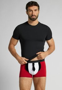 Bóxers rojos con cintura negra y detalle frontal blanco con diseño de corbata negra. El modelo lleva una camiseta ajustada negra.