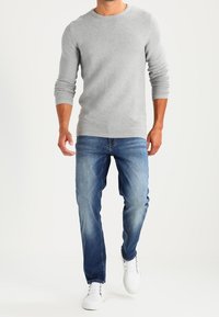 Homme portant un pull texturé gris clair, un jean bleu et des baskets blanches marchant vers l'avant sur un fond blanc.