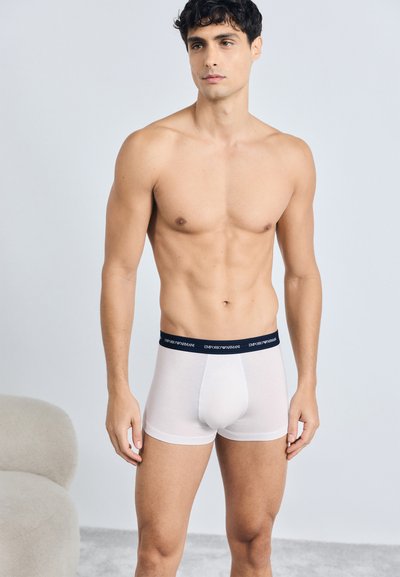 Boxer-briefs en coton blanc avec une ceinture élastique marine portant le nom de la marque "Emporio Armani" en lettres blanches. Texture lisse, design ajusté.