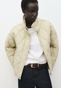 Gequilted crèmekleurige jas met hoge kraag en ritssluiting, gecombineerd met een wit shirt en donkere jeans. Heeft minimale hardware en een gladde textuur.