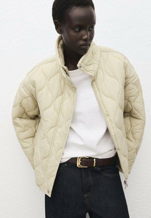 Quilted cream jacket with high collar and zip closure, paired with a white shirt and dark jeans. Features minimal hardware and smooth texture.