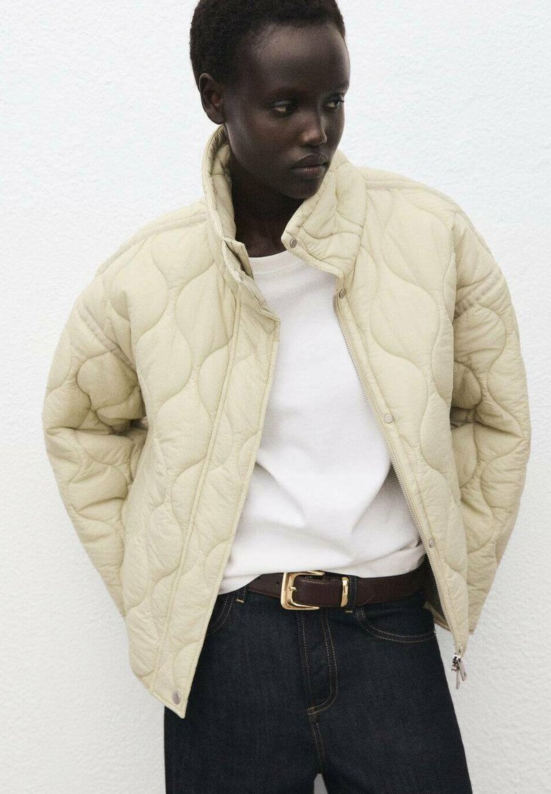 Gequilted crèmekleurige jas met hoge kraag en ritssluiting, gecombineerd met een wit shirt en donkere jeans. Heeft minimale hardware en een gladde textuur.