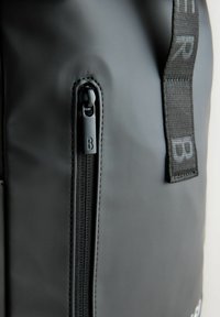 Sac étanche noir avec une surface texturée ; possède une fermeture éclair lisse avec un tirant noir marqué "B" et une sangle à motifs.