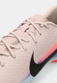 Sapatilha de futebol da Nike em rosa claro com superfície texturizada. Apresenta o swoosh preto, atacadores brancos e realces em degradê de azul e rosa.