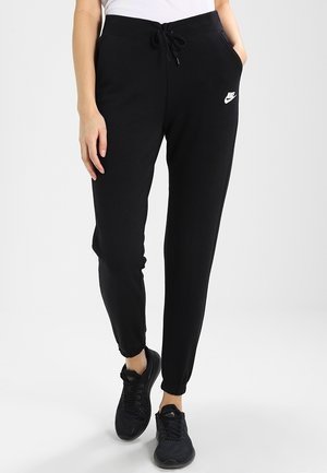 Osoba ubrana w czarne spodnie dresowe Nike z regulowanym paskiem w talii i czarne sportowe buty, stoi z jedną ręką w kieszeni na jednolitym tle.