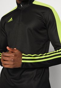 Czarna sportowa bluzka z zapięciem na zamek przy kołnierzu, z jaskrawymi neonowymi akcentami na ramionach i rękawach; zaprojektowana z trzema paskami na każdym rękawie.
