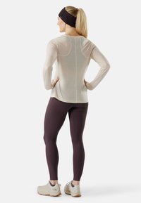 Maglia a maniche lunghe color beige chiaro con texture a rete, leggings aderenti viola scuro e scarpe sportive chiare con suola strutturata.