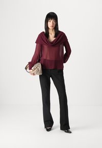 Blusa trasparente borgogna con scollo drappeggiato e maniche lunghe, abbinata a pantaloni neri e una borsa a mano a strisce metalliche.