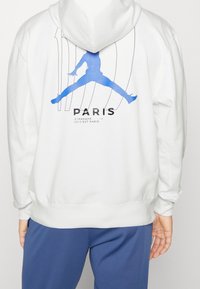 Sudadera blanca con capucha que presenta un gráfico azul de un jugador de baloncesto con el texto "PARÍS" debajo. Textura suave, corte regular y dobladillo acanalado.