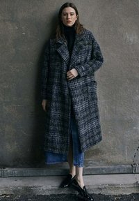 Manteau oversized à carreaux sombres avec un tissu texturé. Présente de grandes poches, un col cranté et une fermeture double boutonnage. Porté avec un jean en denim et des chaussures brillantes.