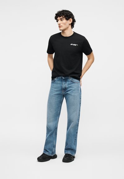 Jeune homme aux cheveux bouclés portant un T-shirt noir à motif, un jean droit bleu clair et des baskets noires, debout les mains derrière le dos.