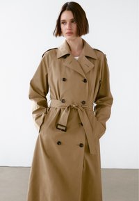 Jasnobrązowy płaszcz trenchowy z podwójnym zapięciem, paskiem w talii i dużymi przednimi guzikami. Posiada klasyczny kołnierz oraz naramienniki.