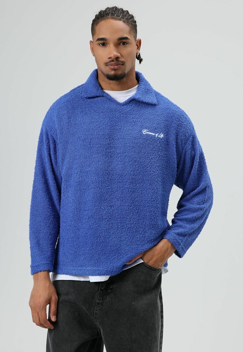 Blauwe fleece trui met kraag, met een gestructureerd oppervlak, lange mouwen en een wit geborduurd logo op de borst. Gestyled met donkere broek.