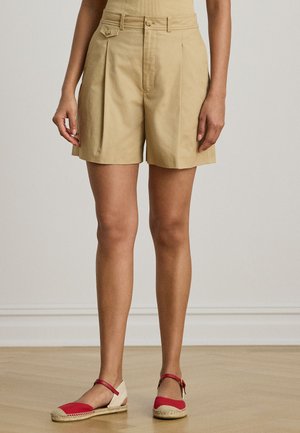 Person iført beige plissert shorts og røde espadrilles med ankelstropper, stående på tregulv mot en ensfarget vegg.