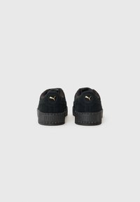 Black suede sneakers with a textured black rubber sole, featuring a gold logo on the back. Low-cut design with padded collar.
