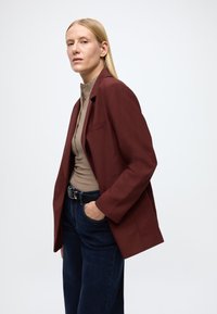 Blazer bordeaux à revers cranté, poche latérale unique, associé à un haut beige côtelé et un jean en denim foncé, mettant en valeur une coupe ajustée.
