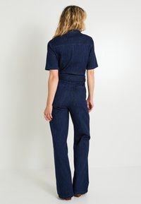 Mujer de pie, de espaldas, vestida con un mono de mezclilla azul oscuro de mangas cortas, perneras anchas y bolsillos de parche en la parte posterior.