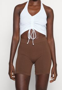 Femme portant un crop top blanc froncé avec cordons et un short cycliste côtelé marron taille haute sur un fond uni.